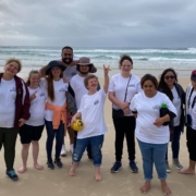 A group of 10 people stand on a beach with arms around each other and smiling.