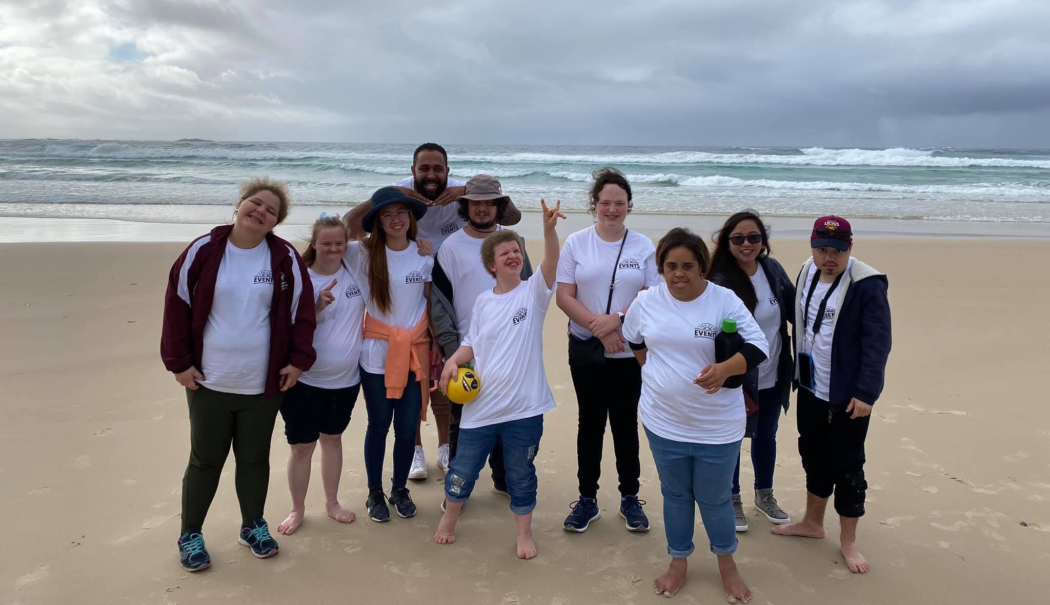 A group of 10 people stand on a beach with arms around each other and smiling.
