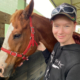 Close shot of a young woman standing next to a horse stroking its jaw. She is smiling at the camera.