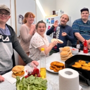 Five young adults stand at a kitchen bench where they are making burgers. There are chicken patties in a frypan, and buns and fresh salad ingredients on the bench. The people are laughing and happy.