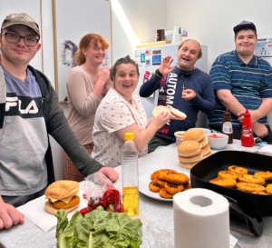 Five young adults stand at a kitchen bench where they are making burgers. There are chicken patties in a frypan, and buns and fresh salad ingredients on the bench. The people are laughing and happy.